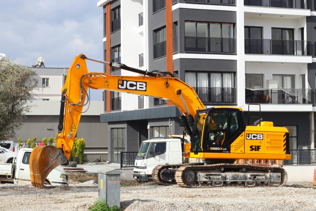 A yellow excavator operating at a city construction site in front of modern buildings.